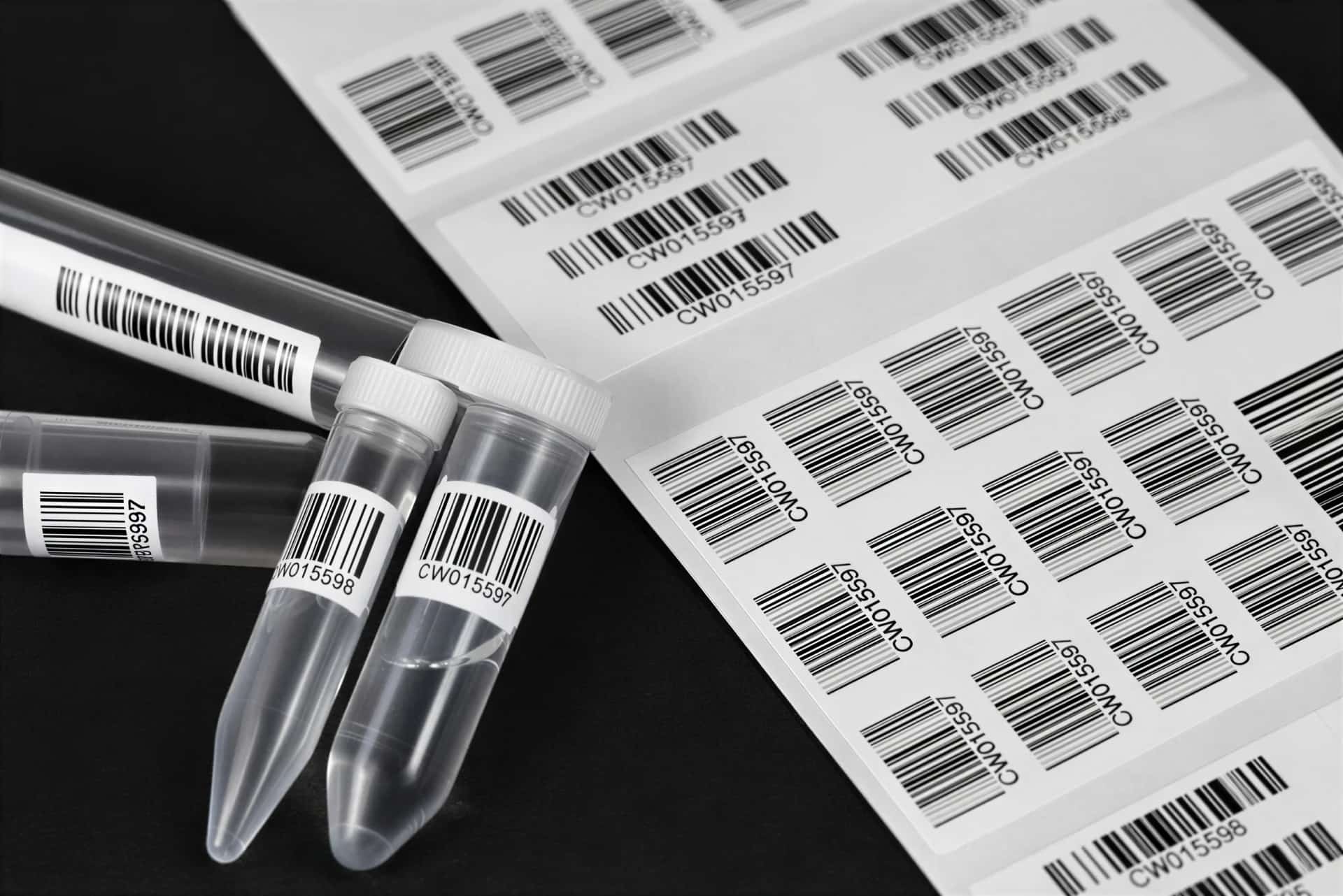 Des tubes d'échantillons étiquetés de codes-barres, posés près de planches d'étiquettes, sur fond noir.